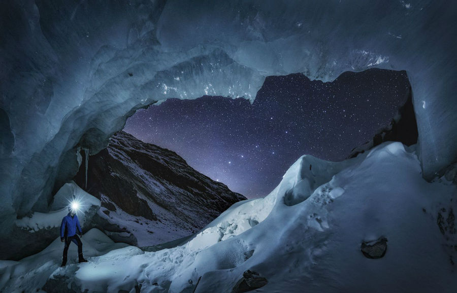 عکاسی که در دروازه غار یخی در انگادین در کشور سوئیس ایستاده و مشغول عکاسی از مجمع الکواکب Big Dipper است.